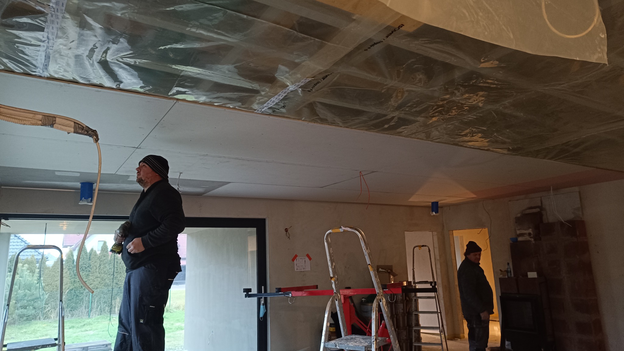 Montaż sufitu podwieszanego z płyty karton gips, widoczne profile, folia izolacyjna i pracownik z wiertarką na drabinie.