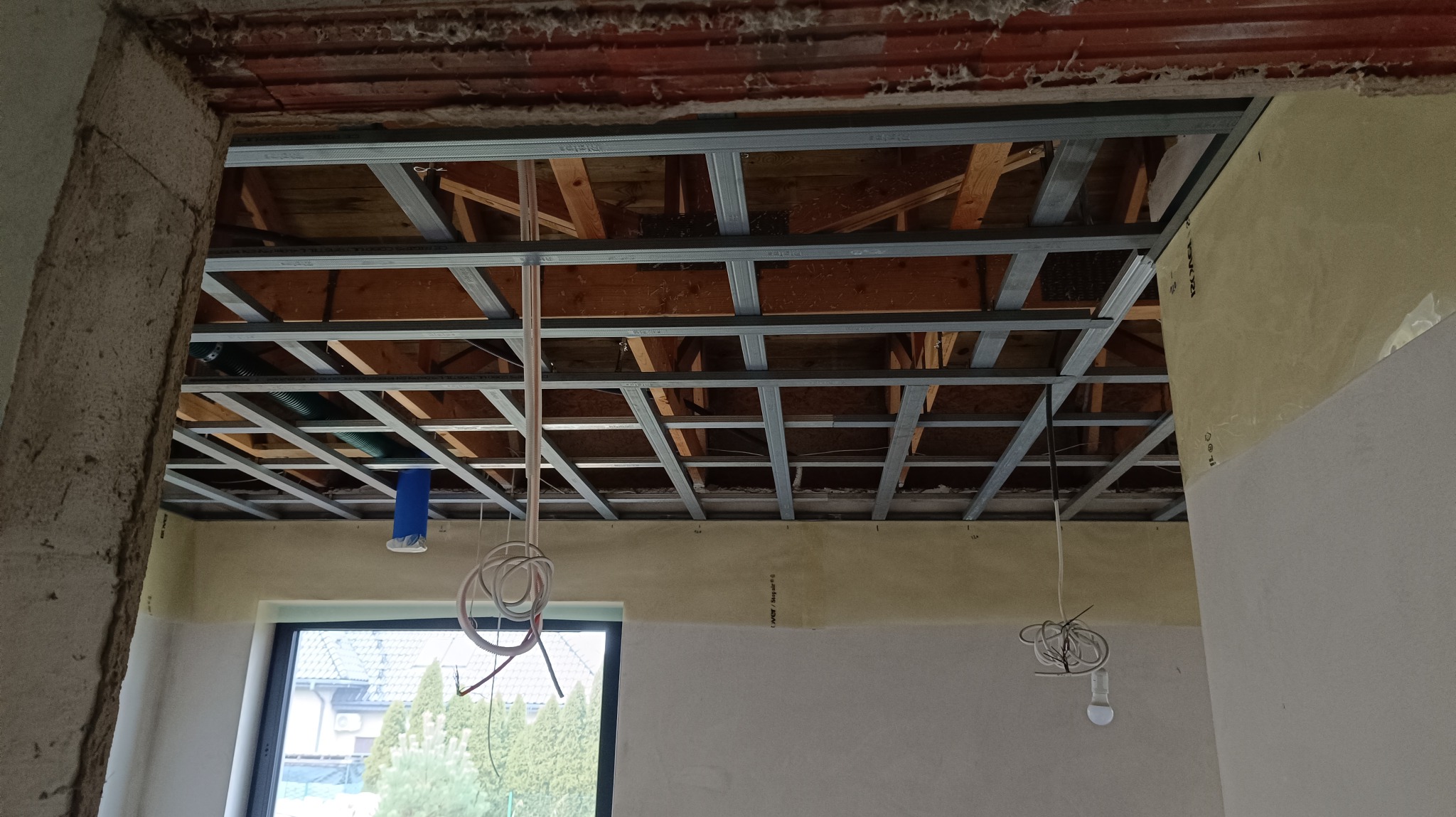 Szkielet sufitu podwieszanego z profili metalowych w trakcie montażu w Lisowicach, widoczne przewody instalacji elektrycznej i fragment okna.