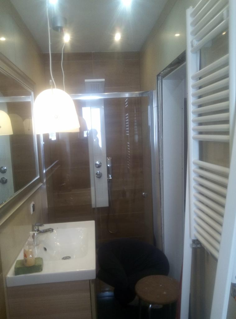 Wnętrze łazienki z kabiną prysznicową wyłożoną brązowymi płytkami, wiszącą lampą z białym kloszem, umywalką z szafką i grzejnikiem drabinkowym; widoczna osoba w ciemnym ubraniu pochylona w kabinie.