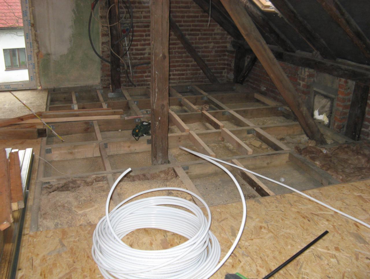 Poddasze w trakcie adaptacji: widoczne drewniane legary pod podłogę, izolacja, przewody elektryczne i rura PCV zwinięta w krąg, pionowy słup konstrukcyjny pośrodku pomieszczenia, fragment okna w tle.