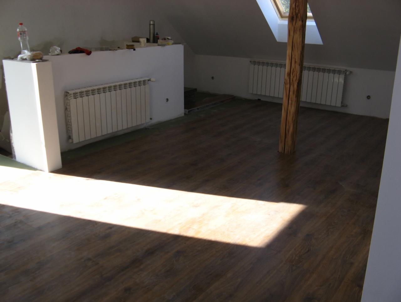 Podłoga z paneli w kolorze ciemnego drewna w pokoju na poddaszu, widoczny słup z surowego drewna, okno dachowe, grzejniki i przygotowane podłoże pod ścianką działową.