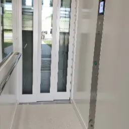 Wnętrze nowoczesnej kabiny osobowej z przesuwnymi drzwiami, panel sterowania ze stali nierdzewnej z podświetlanymi przyciskami, jasna podłoga i oświetlenie sufitowe.