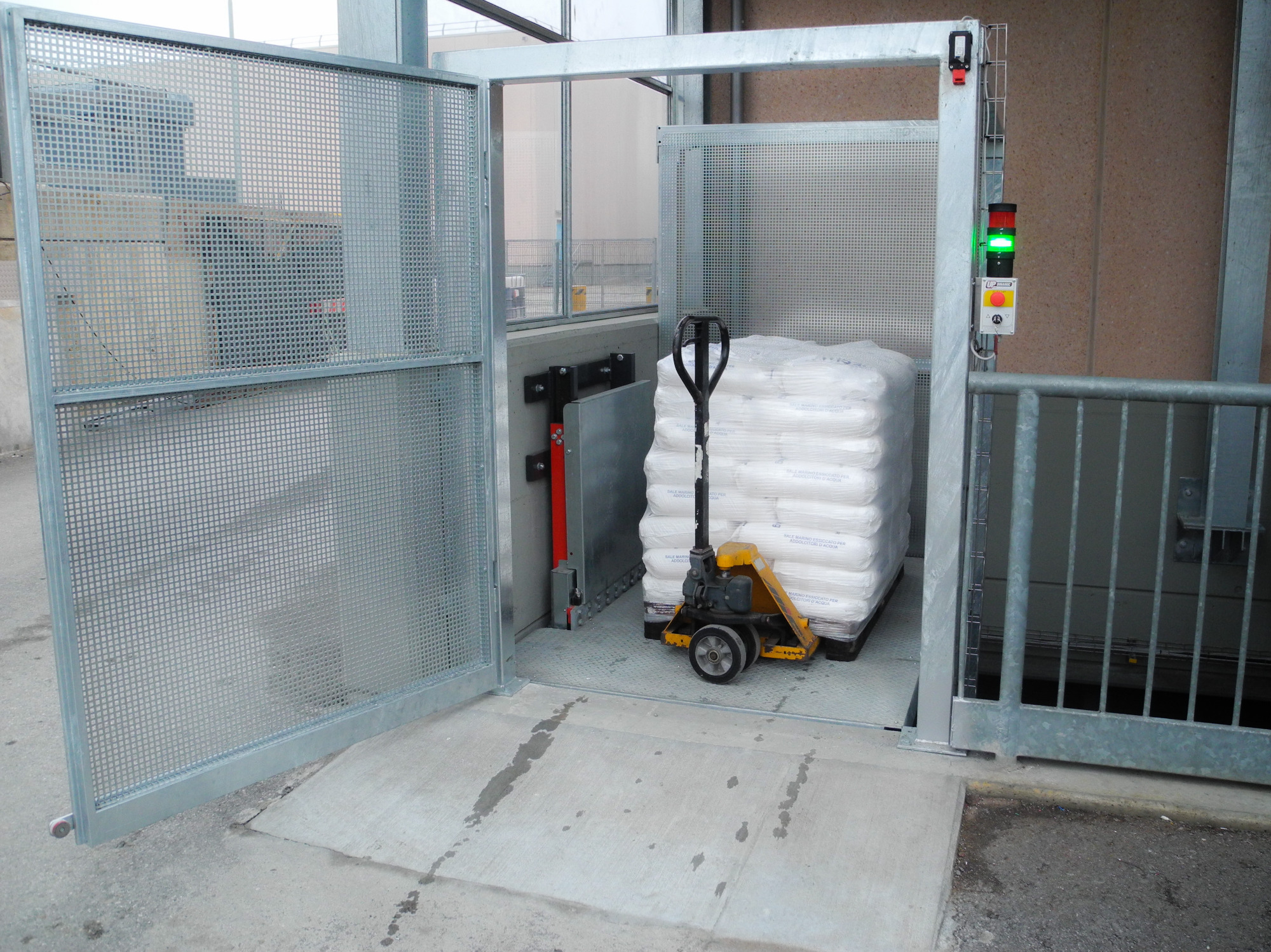 Winda towarowa z otwartymi kratowanymi drzwiami, wewnątrz paleta z workami, obsługa za pomocą wózka paletowego. Widoczny panel sterowania z zieloną lampką.