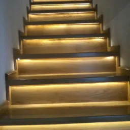 Oświetlone schody z jasnego drewna, widok z dołu, ciepłe światło podkreśla strukturę drewna i krawędzie stopni.