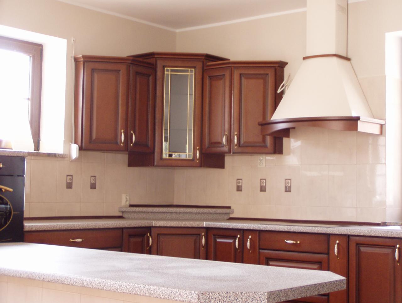 Klasyczna kuchnia z ciemnymi, drewnianymi szafkami, beżowymi płytkami i granitowymi blatami, oświetlona naturalnym światłem z okna.