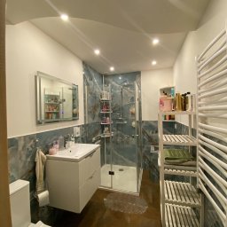 Usługi ogolnobudowlane Józef Nowak - Nowoczesna łazienka z kabiną prysznicową, umywalką z szafką, lustrem z podświetleniem i grzejnikiem drabinkowym, wykończona płytkami imitującymi marmur w odcieniach błękitu i brązu, z brązową...