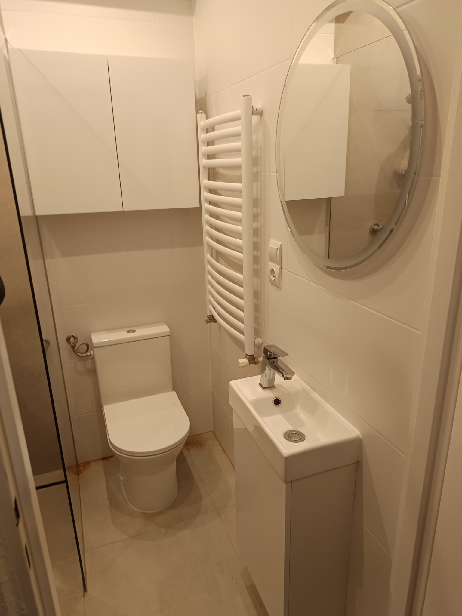 Nowoczesna, biała łazienka z owalnym lustrem bez ramy, grzejnikiem drabinkowym, wiszącą szafką nad toaletą i umywalką z szafką, oświetlona ciepłym światłem.