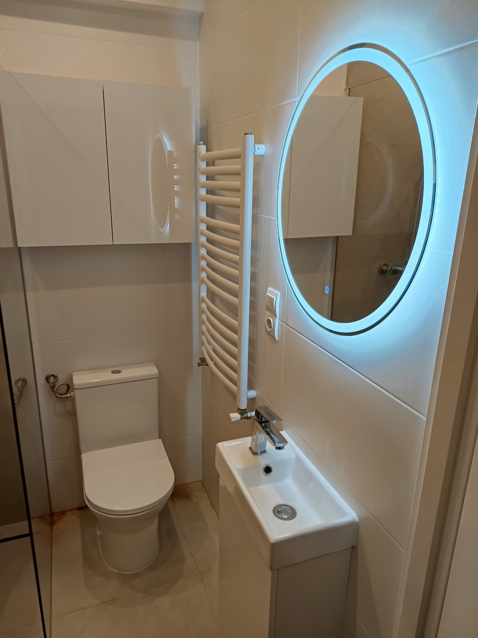 Nowoczesna, jasna łazienka z podświetlanym okrągłym lustrem, wiszącą szafką nad toaletą, grzejnikiem drabinkowym i małą, prostokątną umywalką na szafce.