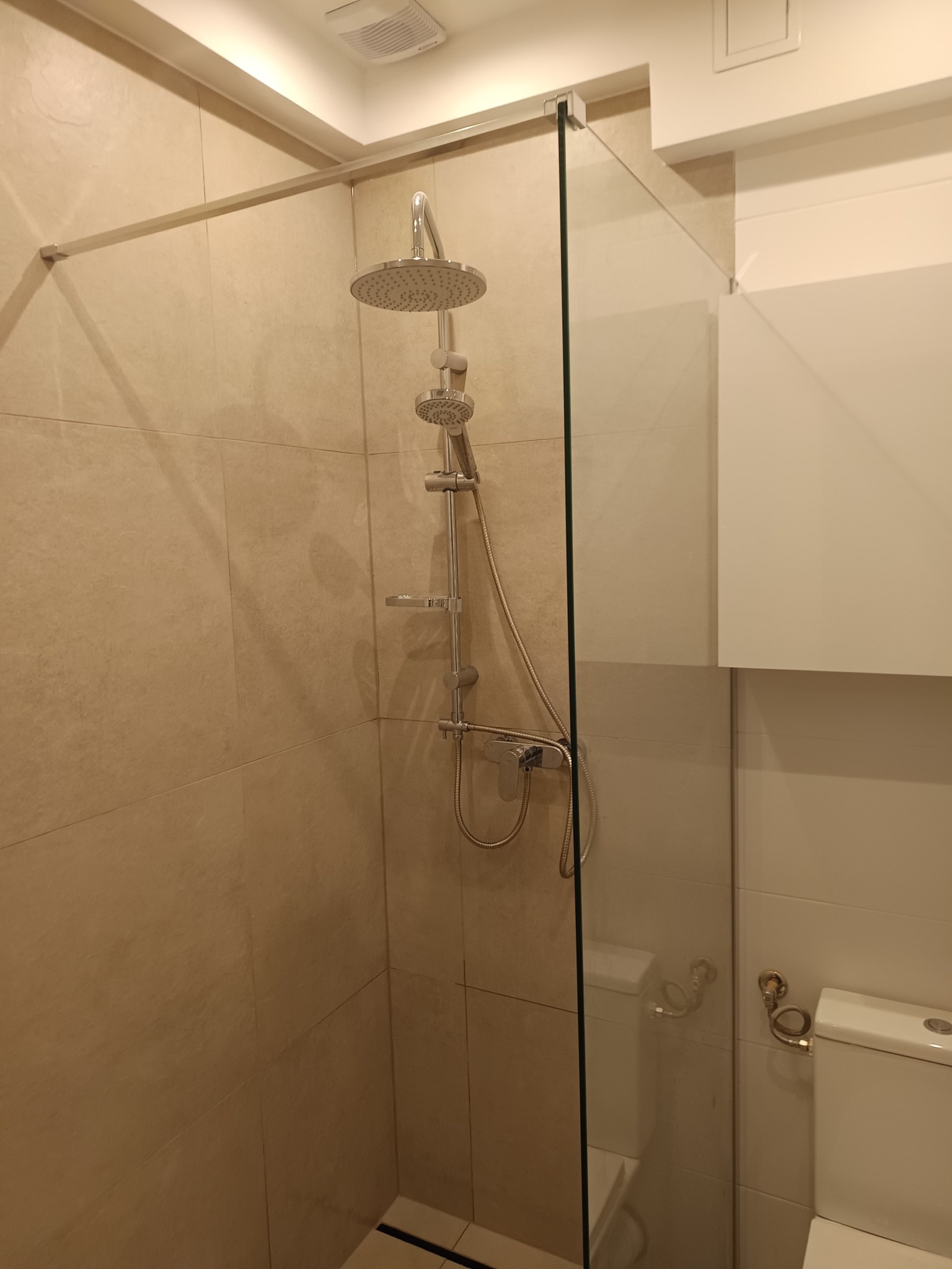 Nowoczesna kabina prysznicowa z deszczownicą i szklaną ścianką, wykończona beżowymi płytkami, z widocznym fragmentem toalety i szafki wiszącej.