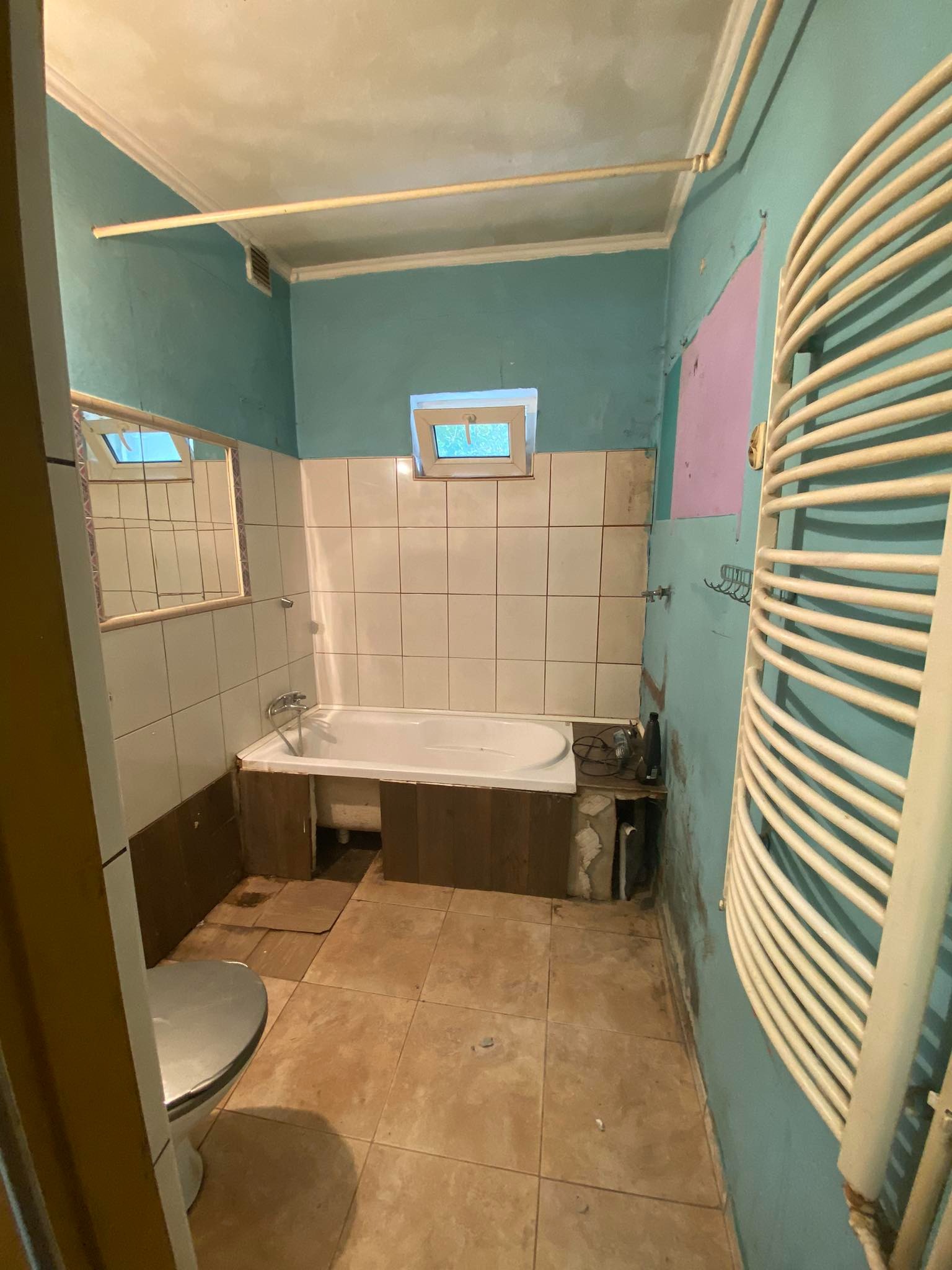 Łazienka w trakcie demontażu: odsłonięte fragmenty ścian, stara wanna obudowana drewnem, grzejnik drabinkowy, brudna podłoga z płytek, toaleta z boku kadru, małe okno nad wanną.