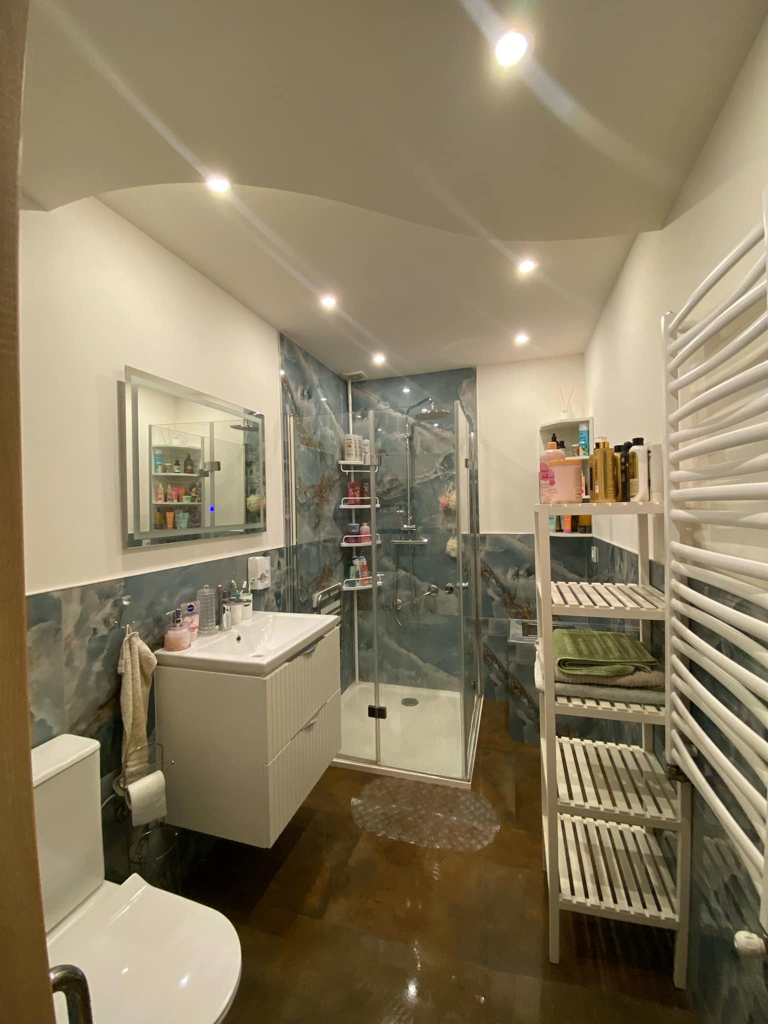 Nowoczesna łazienka z kabiną prysznicową, umywalką z szafką, lustrem z podświetleniem i grzejnikiem drabinkowym, wykończona płytkami imitującymi marmur w odcieniach błękitu i brązu, z brązową...