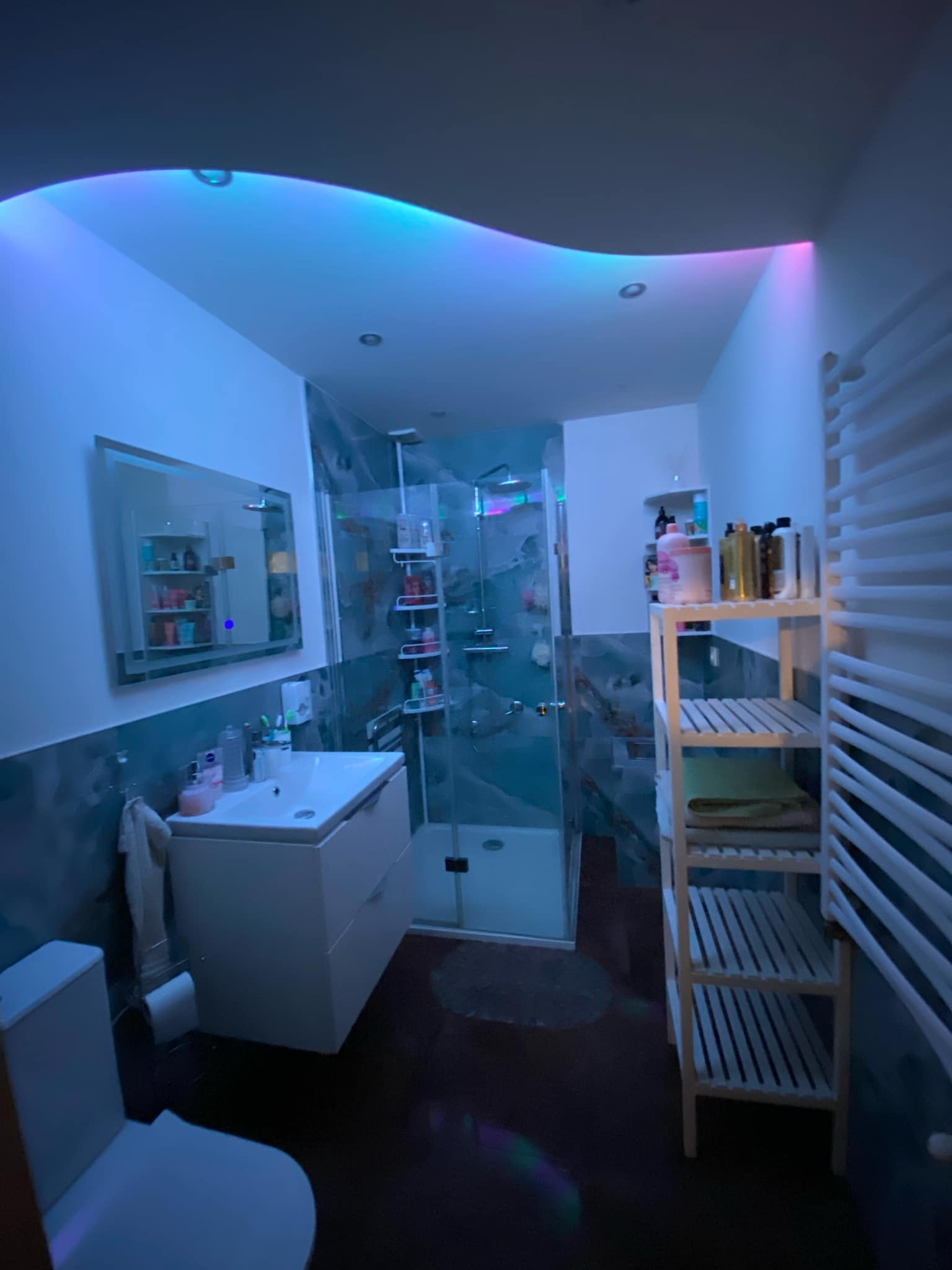 Nowoczesna łazienka z podświetlanym sufitem, kabiną prysznicową ze szklanymi drzwiami, umywalką z szafką, drewnianym regałem na kosmetyki i ręczniki oraz grzejnikiem drabinkowym