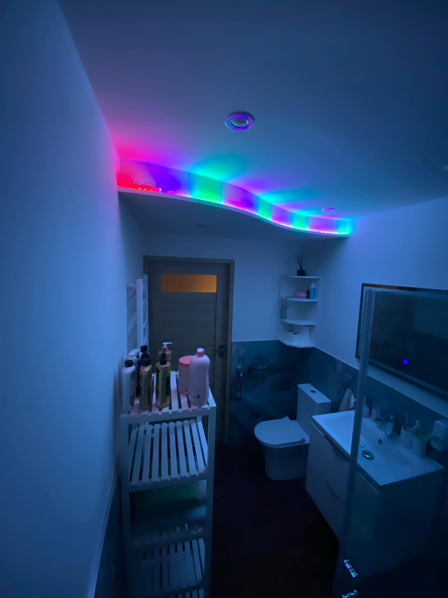 Nowoczesna łazienka z podświetlanym sufitem LED w kolorach tęczy, biała ceramika sanitarna, półki z kosmetykami i akcesoriami, marmurowa ściana.
