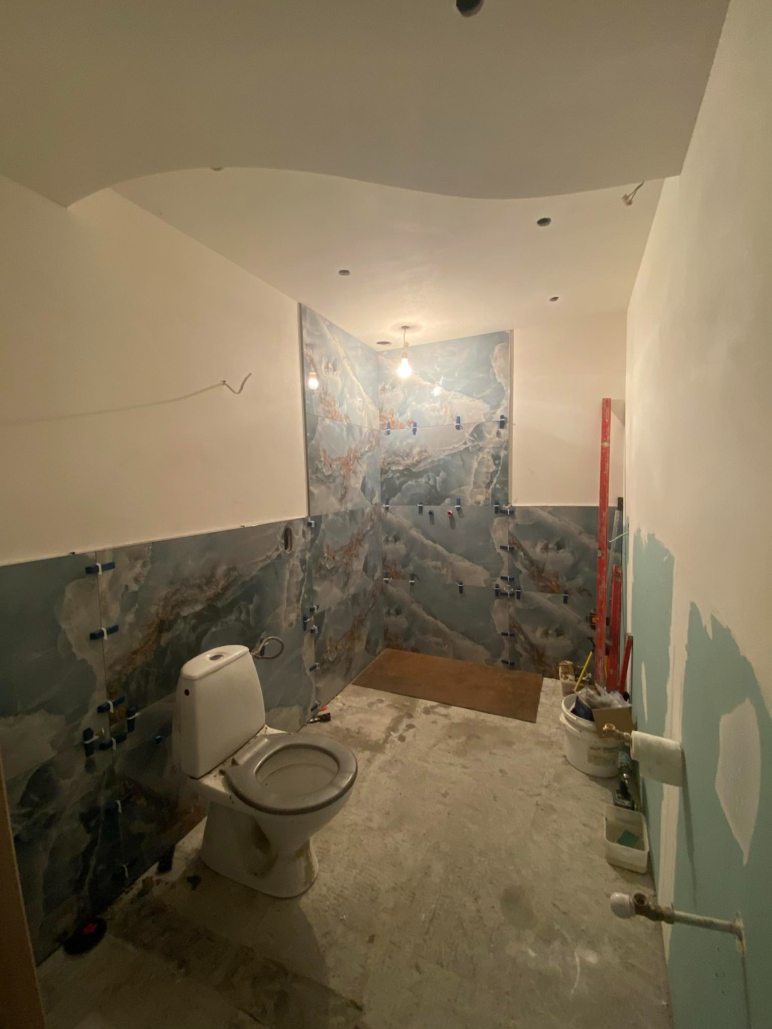Łazienka w trakcie prac wykończeniowych: częściowo ułożone marmurowe płytki na ścianach, wisząca żarówka, niepodłączona toaleta, widoczne narzędzia i akcesoria budowlane.