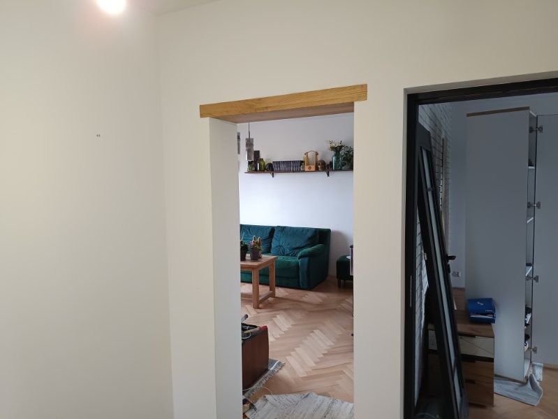 Widok przez odnowione przejście między pokojami, z surową drewnianą belką nad otworem drzwiowym, na salon z parkietem w jodełkę i zieloną sofą.