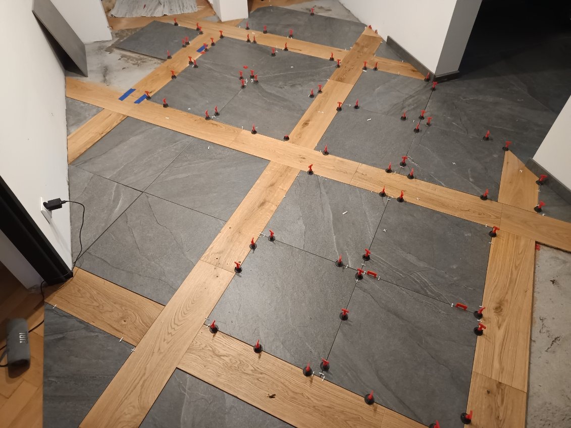 Układanie szarych płytek podłogowych z drewnianymi listwami dekoracyjnymi i czerwonymi klipsami poziomującymi podczas montażu.