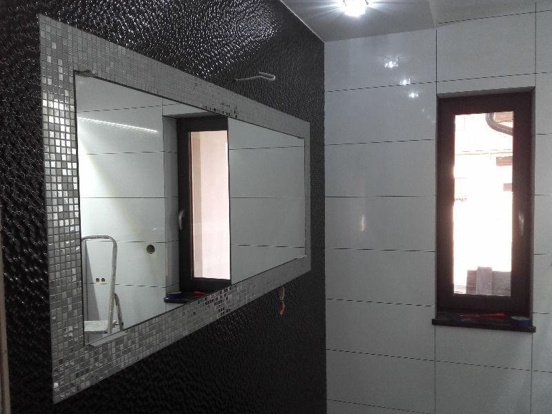 Wykończona łazienka z czarno-białymi płytkami, dużym lustrem z mozaikową ramą i wąskim oknem. W odbiciu lustra widoczny drabinkowy stołek.