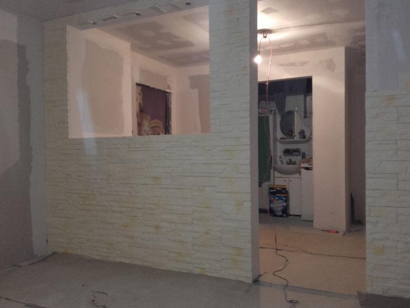 Wnętrze w trakcie wykańczania: ściana z dekoracyjną okładziną imitującą kamień, widok na łazienkę przez otwór w ścianie, surowy strop i podłoga.