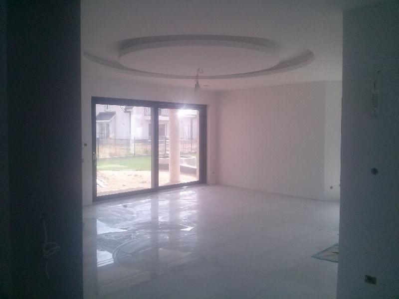 Jasne wnętrze z dużym oknem i błyszczącą, jasną podłogą. Sufit z dekoracyjnym, okrągłym elementem. Widok na ogród przez okno. Minimalistyczny design.