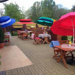 Umbrella - Metalowy grill ogrodowy z drewnianymi elementami, widoczny zza stolików kawiarnianych pod kolorowymi parasolami, na brukowanym patio wśród zieleni.