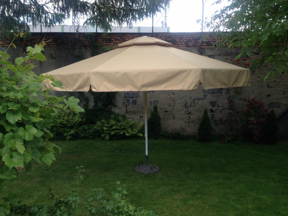 Beżowy parasol ogrodowy z metalową podstawą na trawniku, na tle muru z kamienia i cegły, otoczony zielenią.