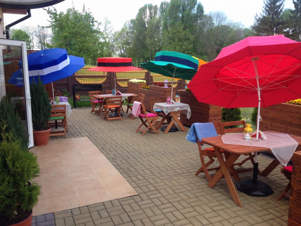 Metalowy grill ogrodowy z drewnianymi elementami, widoczny zza stolików kawiarnianych pod kolorowymi parasolami, na brukowanym patio wśród zieleni.