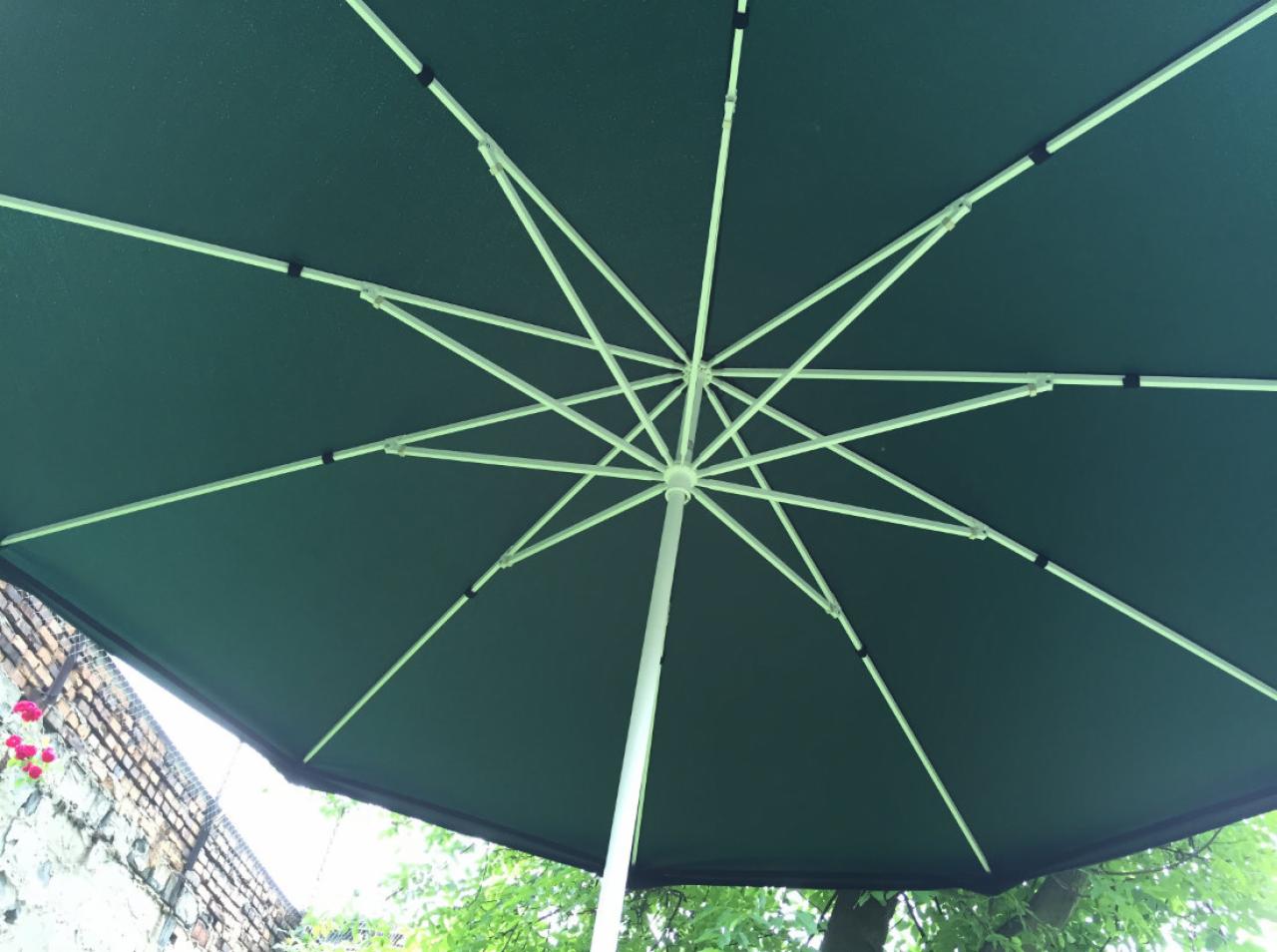 Widok od dołu na zielony parasol ogrodowy z białym stelażem, na tle muru i zieleni. Konstrukcja metalowa z elementami mocującymi.