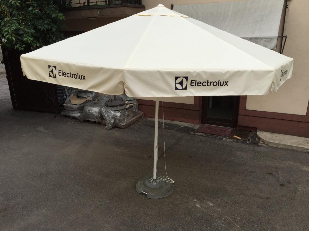 Biały parasol ogrodowy z logo Electrolux na szarym podłożu, na zewnątrz. W tle widoczne elementy metalowe i fragment budynku.