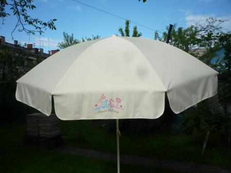Jasny parasol ogrodowy z dziecięcym motywem, widoczny na tle zielonego ogrodu i budynku. Charakterystyczny wzór na materiale. Oświetlenie dzienne.
