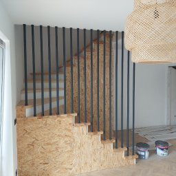 CORINA - Schody z płyty OSB z czarnymi, pionowymi balustradami, widoczne puszki z farbą na podłodze, w tle wisząca, pleciona lampa i otwarte okno.