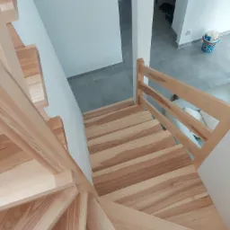 Widok z góry na jasne, drewniane schody z poręczą, skręcające w dół; szare płytki na podłodze w tle, z widocznymi butami i akcesoriami w przedpokoju.