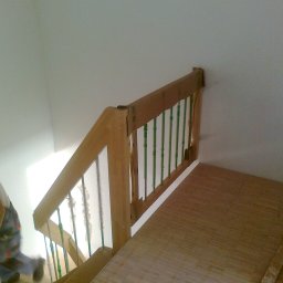 CORINA - Fragment schodów z drewnianą balustradą z zielonymi prętami, w trakcie montażu, widoczne niedociągnięcia i zabezpieczenia na prętach, podłoga wyłożona panelami.