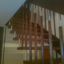 CORINA - Klatka schodowa z drewnianymi schodami i metalowymi balustradami, widok z dołu, ściana w pionowe pasy w tle.