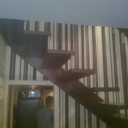 CORINA - Schody drewniane wspornikowe z metalowymi elementami mocującymi, widoczne na tle ściany w pionowe pasy, z prześwitem do innego pomieszczenia w dolnej części kadru.