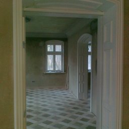 CORINA - Wnętrze pokoju w trakcie remontu, widok przez białe, łukowe drzwi, z wzorzystą podłogą i oknem w tle.