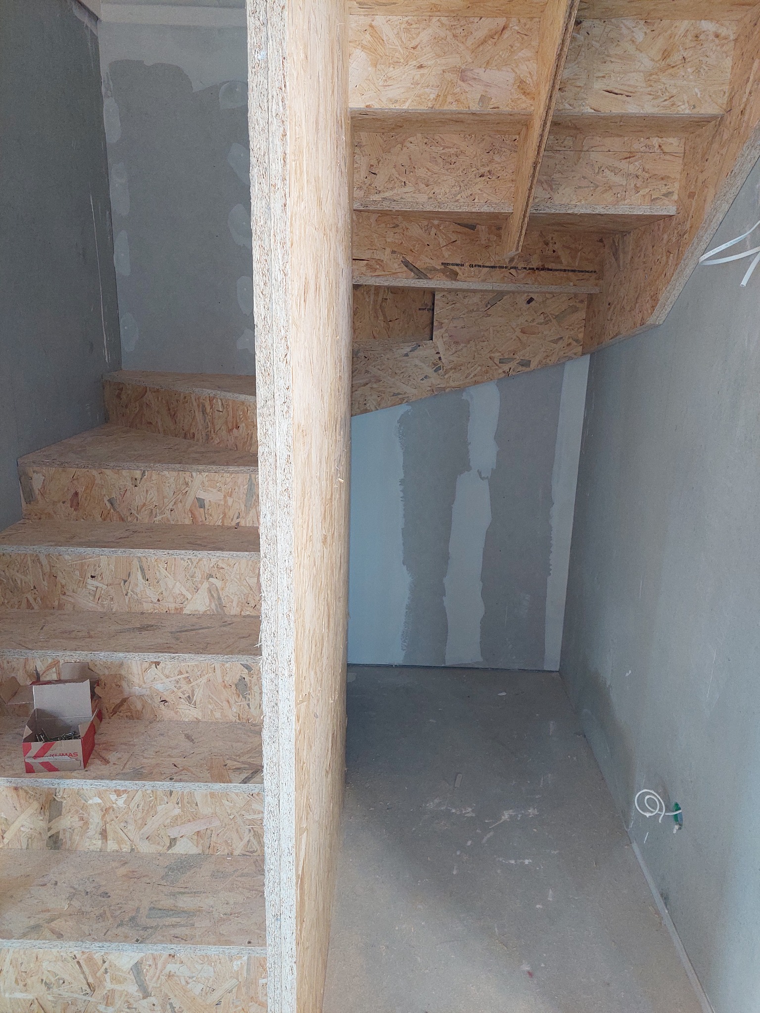 Surowe, drewniane schody z płyty OSB w trakcie budowy, widoczna puszka z wkrętami na jednym ze stopni, ściany w stanie surowym.