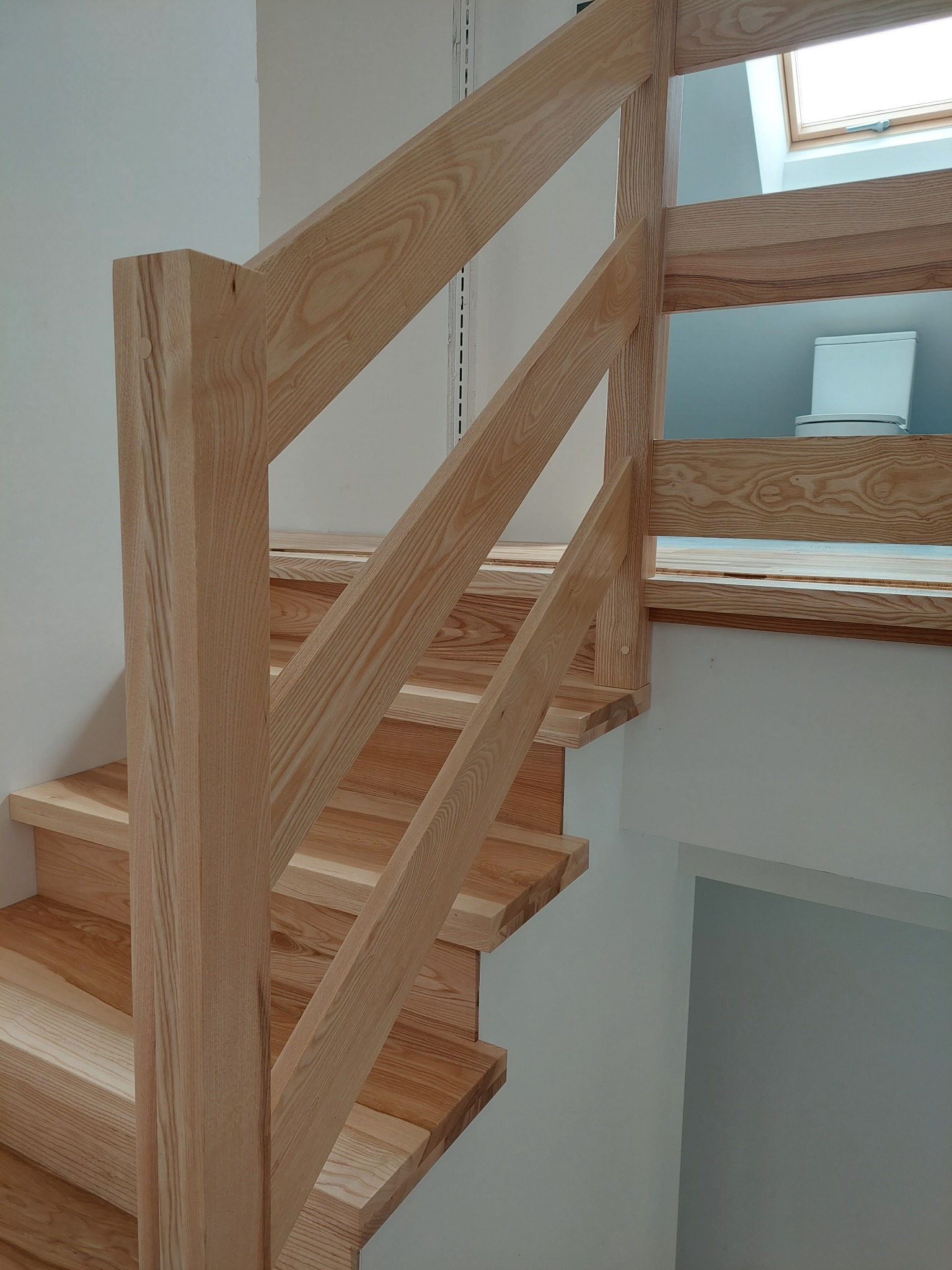 Jasne drewniane schody z balustradą o prostym designie, widok z dołu, w tle biała ściana i okno dachowe z widoczną toaletą.