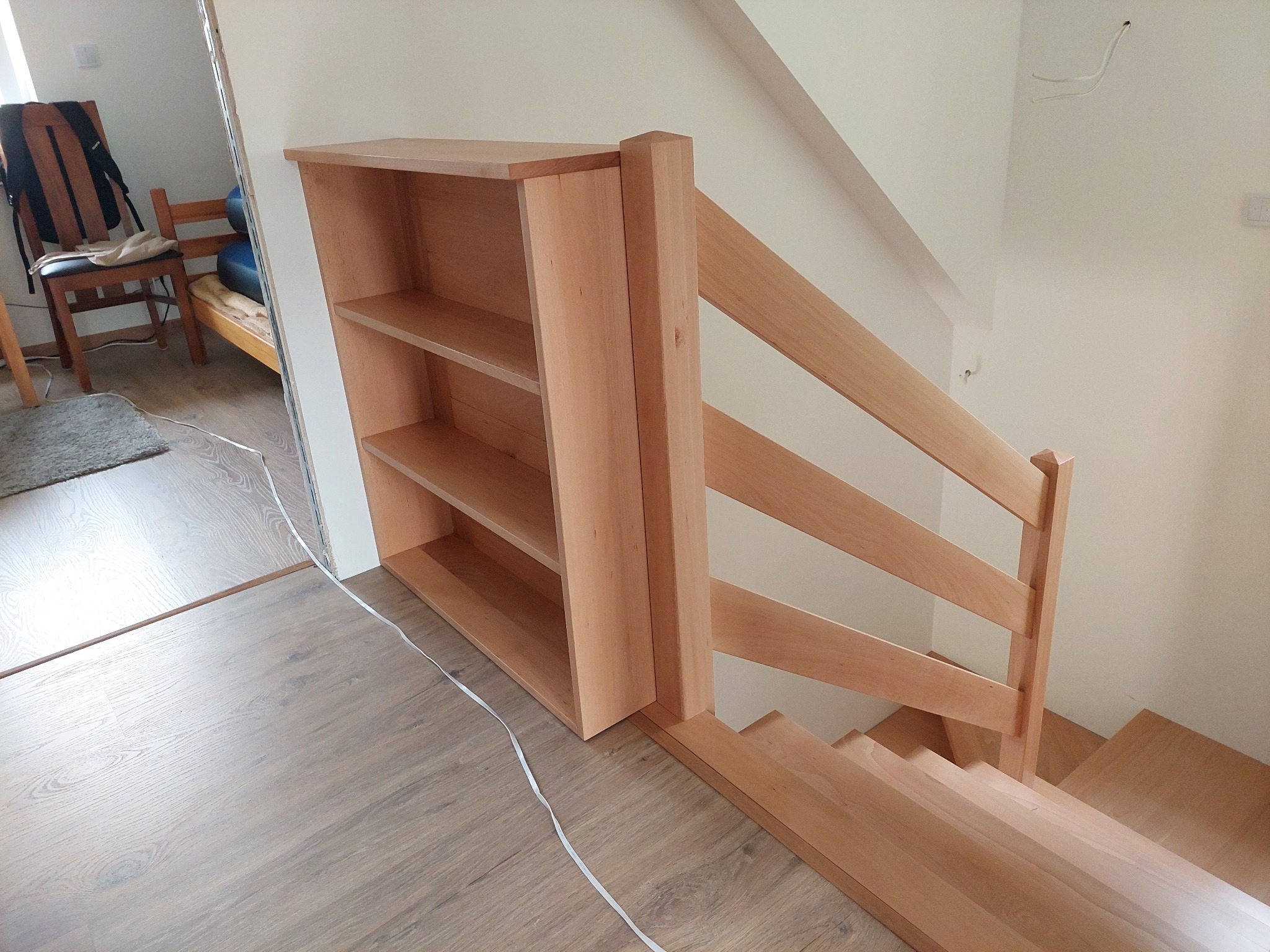 Jasne, drewniane schody z barierką, zintegrowane z regałem na książki, widoczne na tle białej ściany i podłogi z paneli.