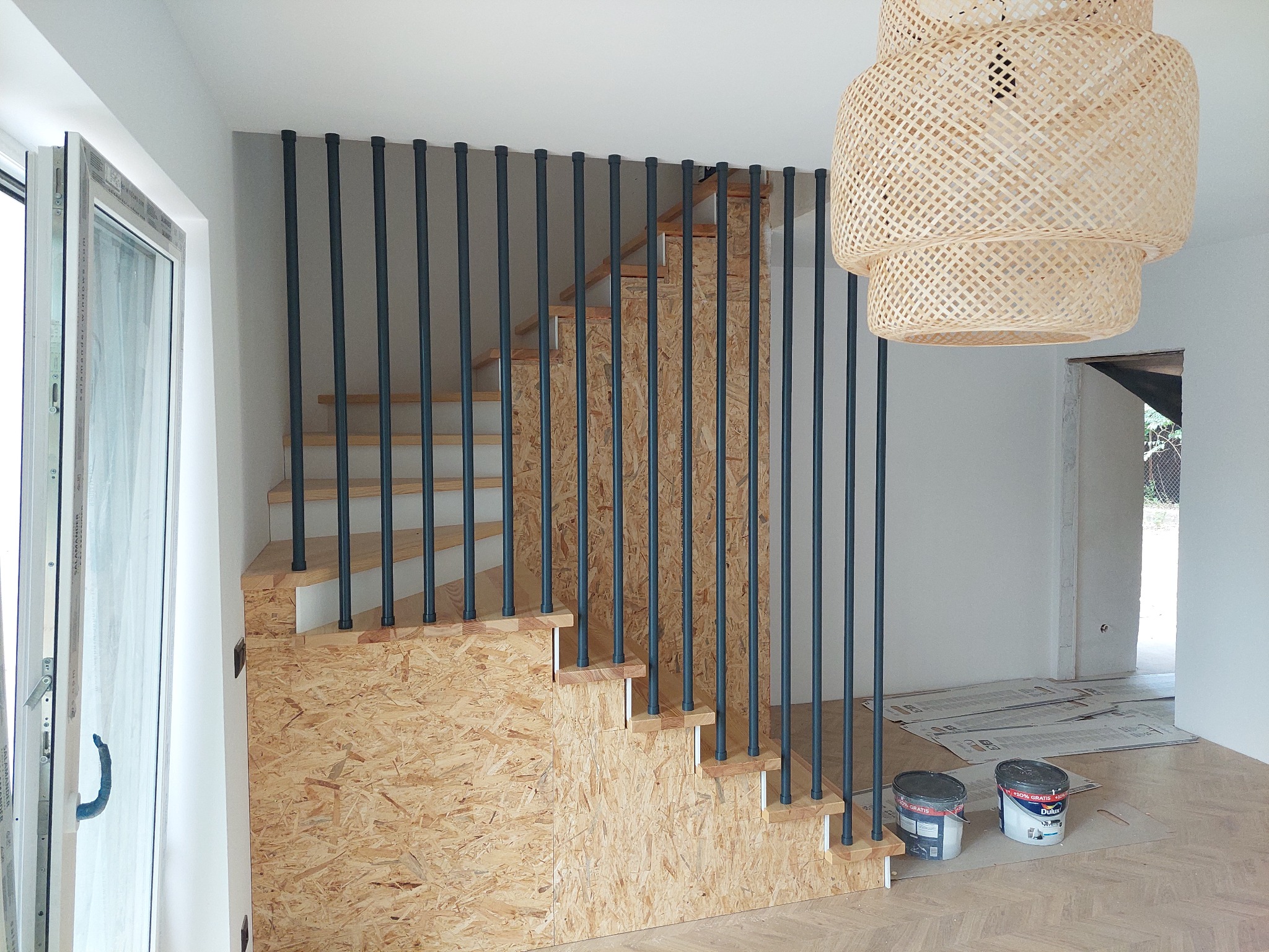 Schody z płyty OSB z czarnymi, pionowymi balustradami, widoczne puszki z farbą na podłodze, w tle wisząca, pleciona lampa i otwarte okno.