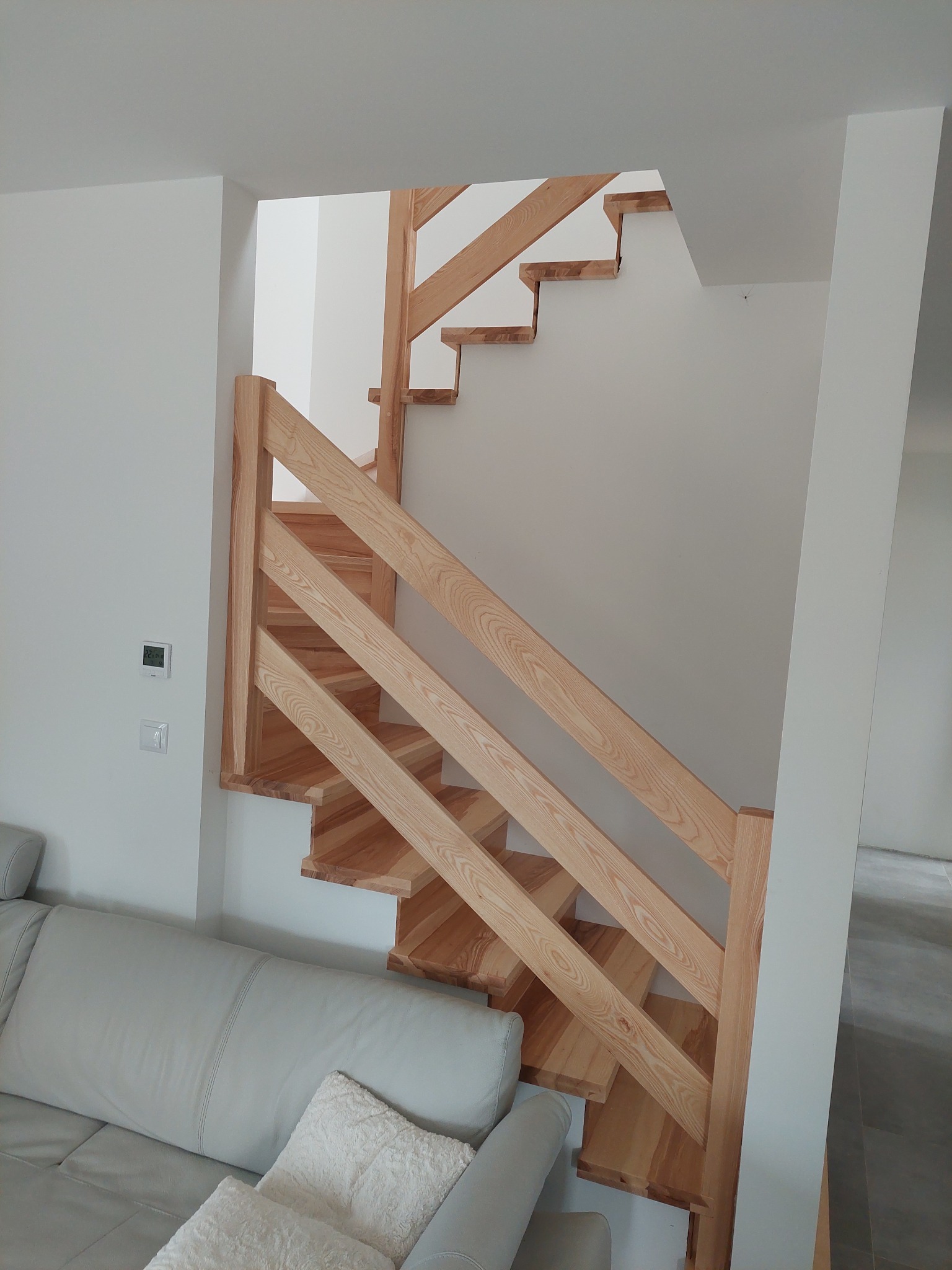 Jasne, drewniane schody z prostymi balustradami w nowoczesnym wnętrzu, widoczne z perspektywy niskiego kąta, obok fragment sofy.