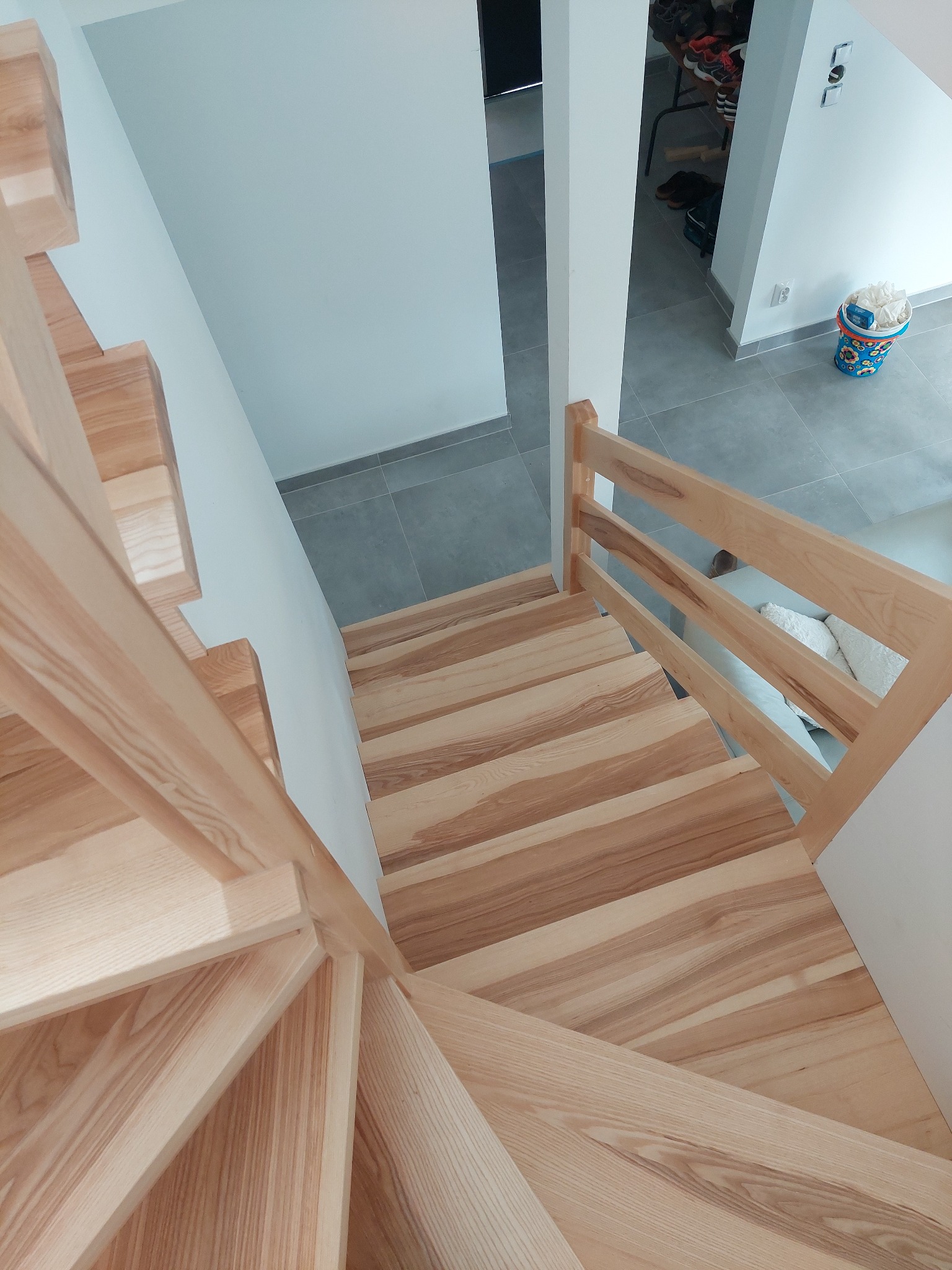 Widok z góry na jasne, drewniane schody z poręczą, skręcające w dół; szare płytki na podłodze w tle, z widocznymi butami i akcesoriami w przedpokoju.