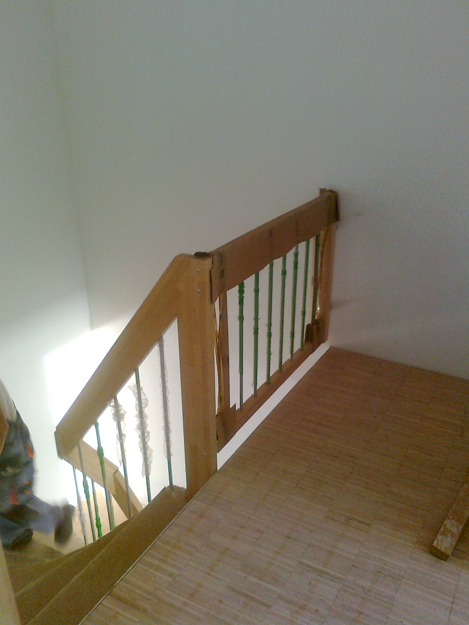 Fragment schodów z drewnianą balustradą z zielonymi prętami, w trakcie montażu, widoczne niedociągnięcia i zabezpieczenia na prętach, podłoga wyłożona panelami.