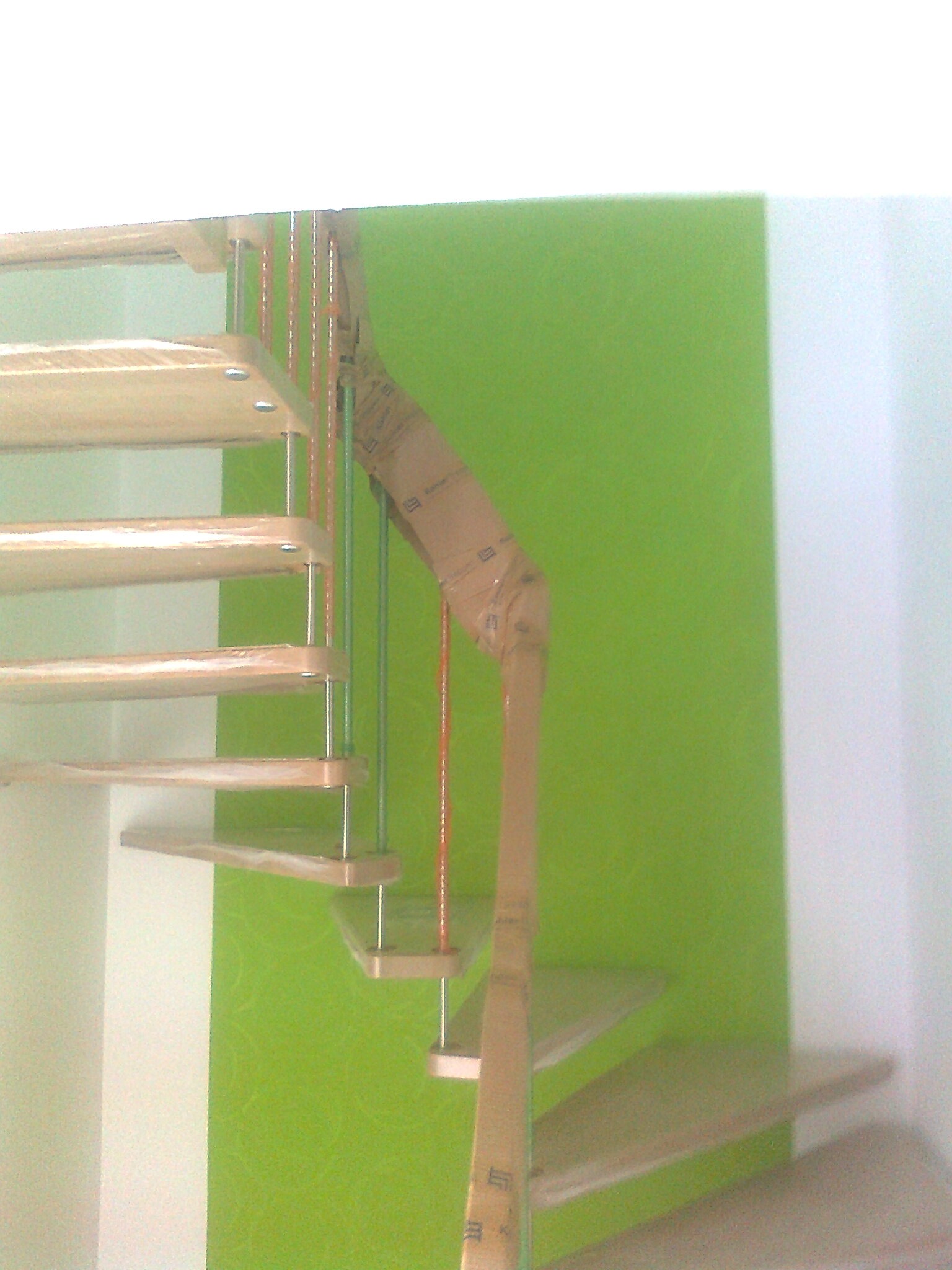 Drewniane schody wiszące z metalowymi wspornikami, zabezpieczone folią ochronną, na tle zielonej ściany.