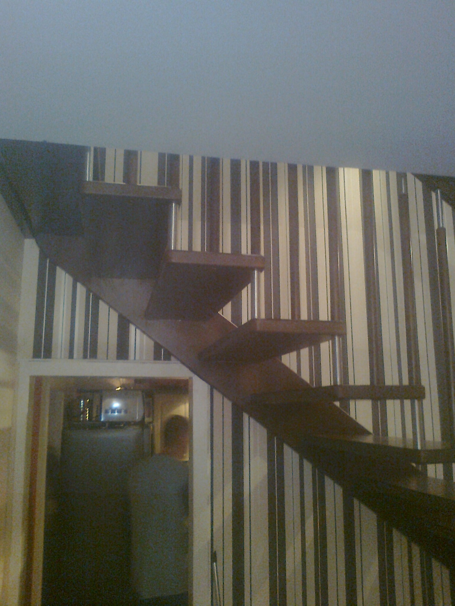 Schody drewniane wspornikowe z metalowymi elementami mocującymi, widoczne na tle ściany w pionowe pasy, z prześwitem do innego pomieszczenia w dolnej części kadru.