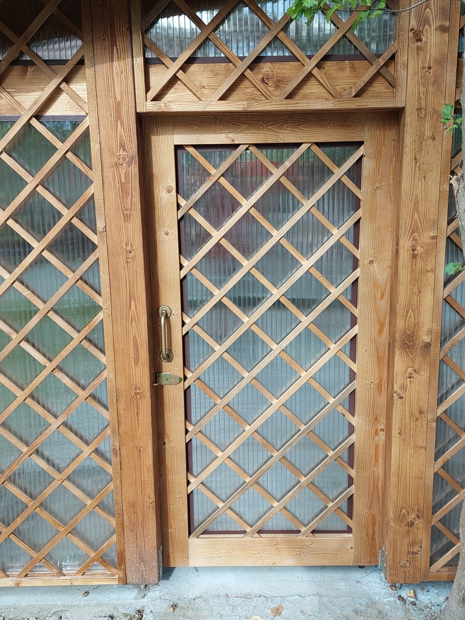 Drewniana konstrukcja z kratownicą i przezroczystym wypełnieniem, prawdopodobnie boczna ściana z drzwiami, z widocznymi słojami drewna i metalową klamką.