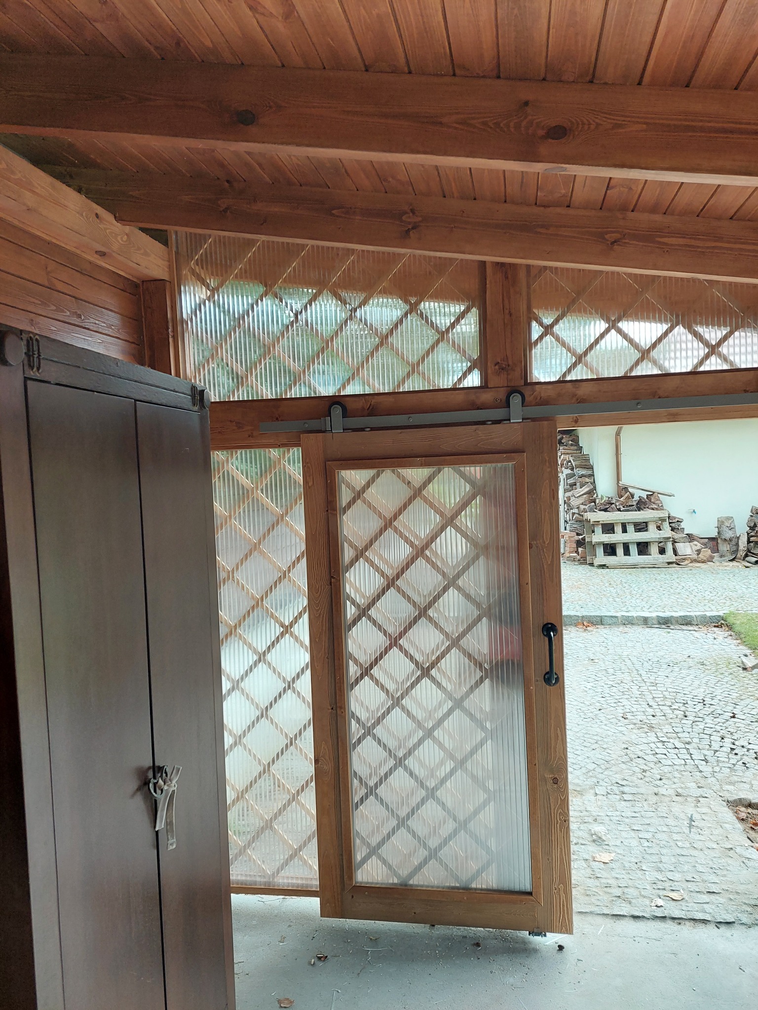 Drewniana konstrukcja z przesuwnymi drzwiami z przezroczystym wypełnieniem w kratkę, widok na stertę drewna i brukowany chodnik.