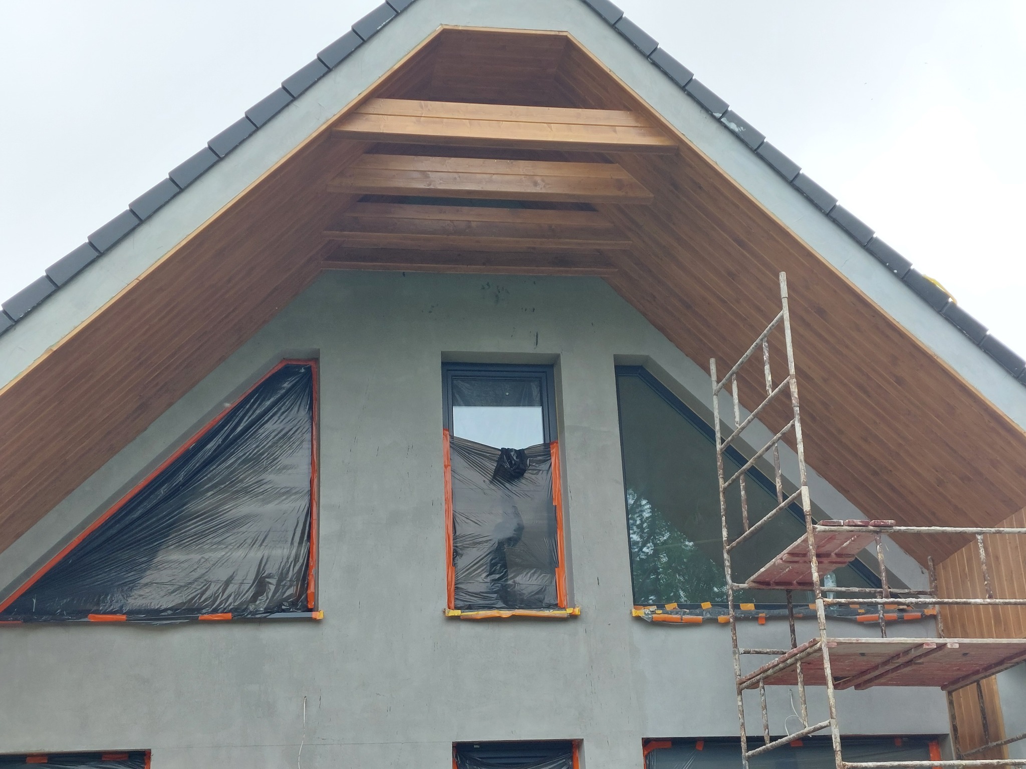 Surowy stan elewacji domu z drewnianym podbitką i zabezpieczonymi folią oknami, widoczne rusztowanie.