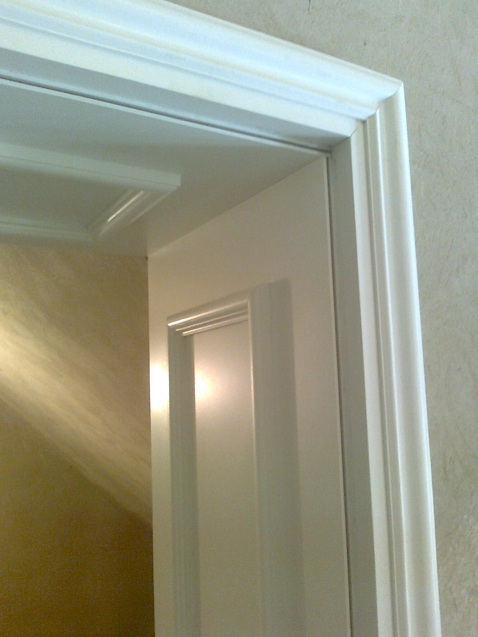 Biała ościeżnica i fragment drzwi wewnątrz pomieszczenia, widoczne detale wykończenia i faktura ściany.