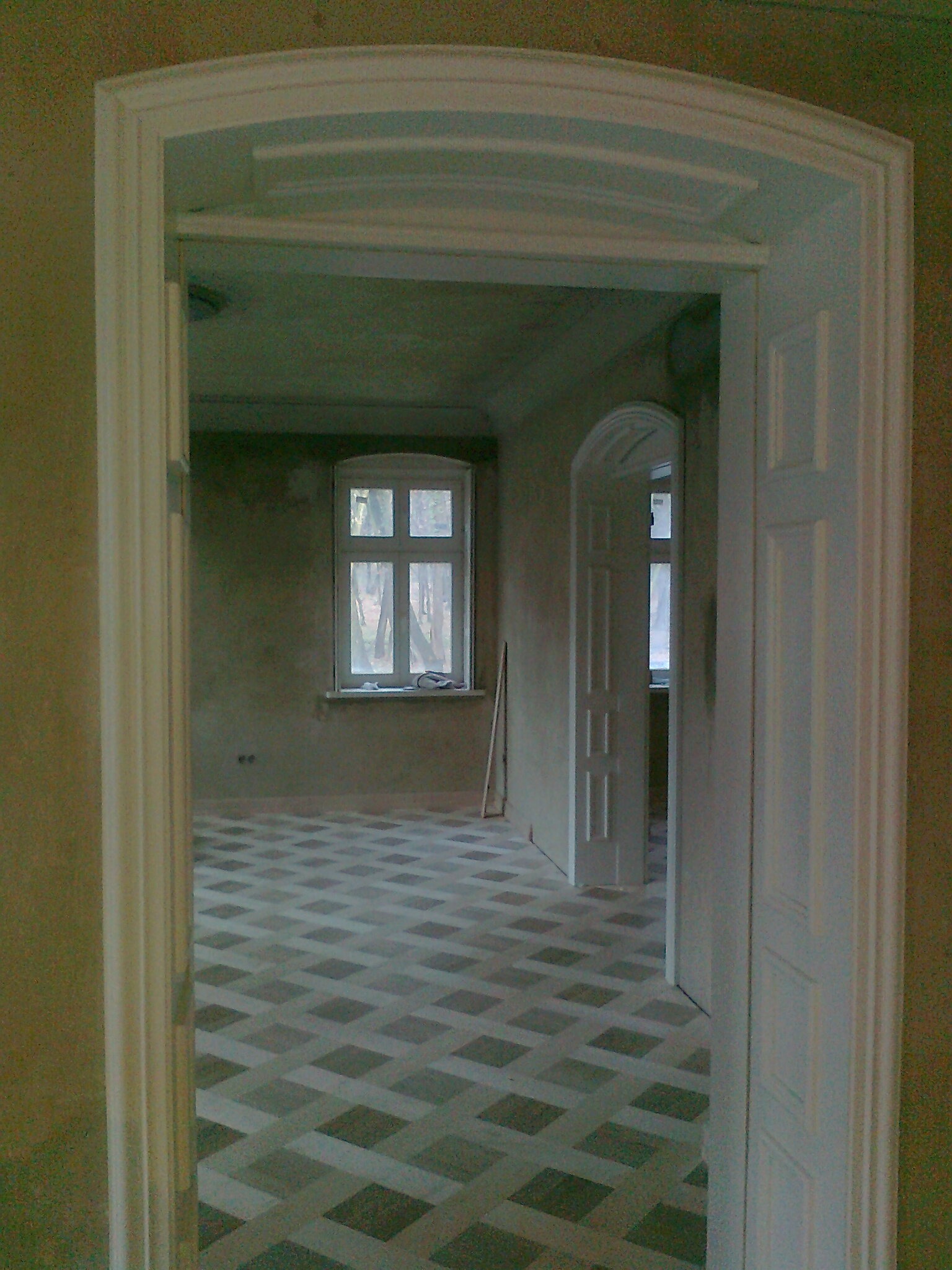 Wnętrze pokoju w trakcie remontu, widok przez białe, łukowe drzwi, z wzorzystą podłogą i oknem w tle.