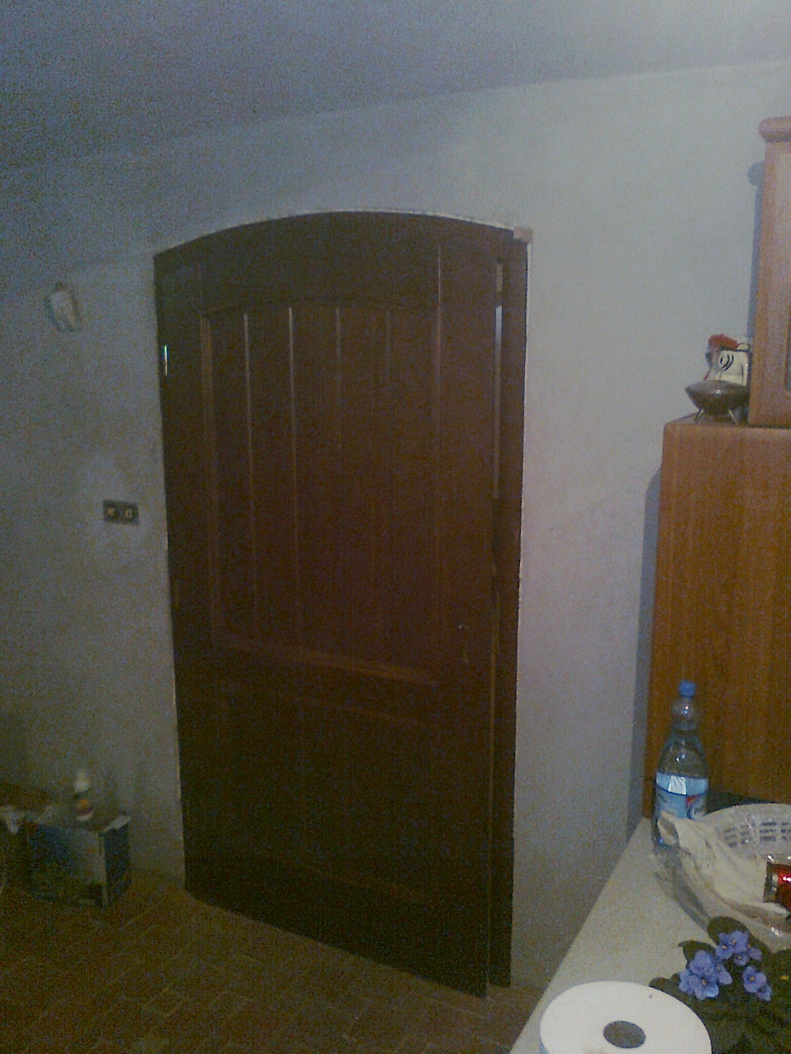 Ciemnobrązowe, drewniane drzwi wewnętrzne z zaokrągloną górą, częściowo otwarte, widoczne na tle ściany z włącznikiem elektrycznym, obok szafki z dekoracją i butelką wody.